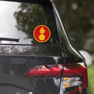 May include: A round sticker featuring a yellow and red design. The sticker has a red background with a yellow circle in the center. A yellow flame design sits atop the yellow circle. The sticker is affixed to the rear window of a black vehicle.