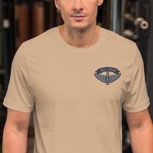 May include: A tan t-shirt featuring a small emblem on the upper left chest. The emblem includes the text "160th SOAR" above a design of wings and a sword. The shirt is made of a soft material and has a crew neck.