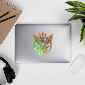 May include: A holographic sticker featuring a crest design with an eagle, trident, and anchor. The sticker has a gradient of colors, including green, orange, and yellow. The sticker is on a laptop.