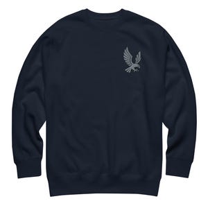 Può includere: Una felpa blu navy a maniche lunghe con un piccolo ricamo grigio raffigurante un'aquila con le ali spiegate, posizionato sulla parte superiore sinistra del petto. Il capo presenta un girocollo, polsini e orlo a coste.