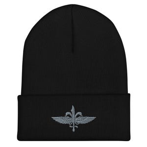 May include: Black beanie with a gray embroidered emblem featuring a stylized fleur-de-lis with wings. The beanie is made of a soft, knitted material and has a folded brim. A stylish and warm accessory for cold weather.