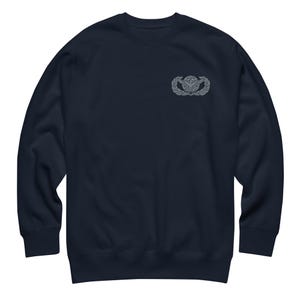May include: A navy blue long-sleeve sweatshirt featuring a small, embroidered grey emblem on the upper left chest. The emblem depicts a stylized eagle with wings spread over a globe, framed by a circular wreath design, centered on the fabric.