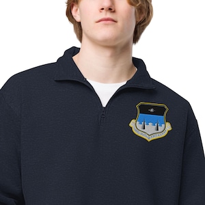 May include: Navy blue quarter-zip sweatshirt with a detailed embroidered patch. The patch features a shield design with blue, black, and yellow accents, and the text "UNITED STATES AIR FORCE ACADEMY".