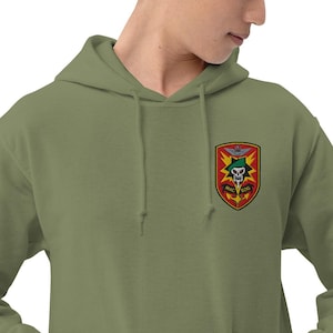 Könnte beinhalten: Olivgrüner Kapuzenpullover mit einem Aufnäher auf der Brust. Der Aufnäher zeigt einen Totenkopf, Flügel und den Text "MAC SOG" in einem Schilddesign. Der Kapuzenpullover hat eine Kapuze mit Kordelzug und eine Kängurutasche.