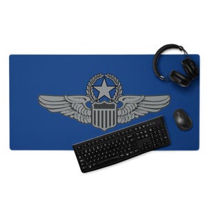 Puede incluir: Una alfombrilla de escritorio azul con un emblema gris de estrella, alas y escudo. Un teclado, un ratón y unos auriculares negros están sobre la alfombrilla. La alfombrilla de escritorio está diseñada para una configuración informática.