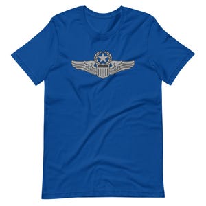 Pode incluir: T-shirt azul royal com um emblema prateado que apresenta asas, uma estrela e um escudo. A t-shirt de manga curta é feita de um material macio, adequada para roupa casual.