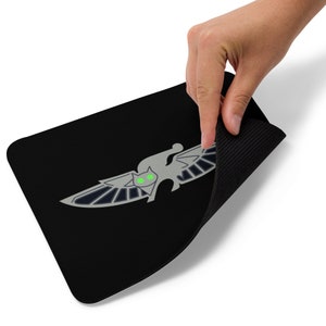 May include: Black mouse pad with a grey winged cat design with green eyes. The mouse pad is rolled up and being held by a hand.
