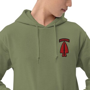 Pode incluir: Moletom com capuz verde-oliva com um patch vermelho e preto que diz "AIRBORNE" acima de uma seta vermelha. O moletom tem um capuz com cordão e um bolso canguru.