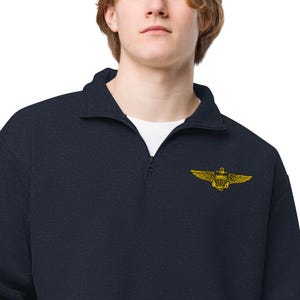 May include: Navy blue quarter-zip sweatshirt with a gold-colored pilot's wings emblem on the chest. The sweatshirt has a ribbed collar and a white undershirt is visible. The emblem features a shield, wings, and a propeller.