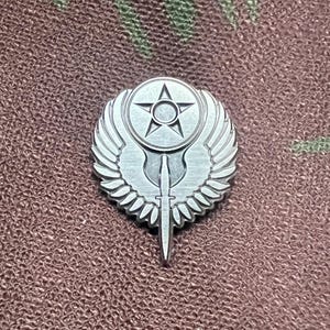 May include: A silver-colored metal pin featuring a star within a circle, wings, and a dagger. The pin is detailed with etched lines and is set against a camouflage background.