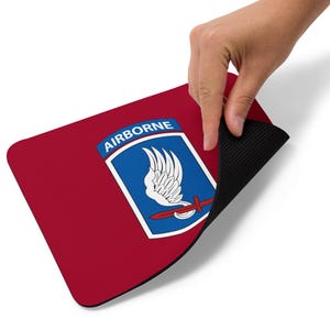 Könnte beinhalten: Ein rotes rechteckiges Mauspad mit schwarzer Unterseite. Das Mauspad zeigt ein blau-weißes Emblem mit dem Wort "AIRBORNE" oben. Das Emblem enthält einen weißen Flügel, ein rotes Schwert und einen blauen Hintergrund.