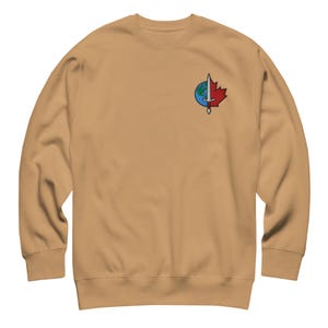 May include: A tan crewneck sweatshirt featuring a small embroidered logo on the upper left chest. The logo depicts a vertical silver sword centered over a split circular design, with a blue and green globe on the left and a red maple leaf on the right.