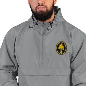 Pode incluir: Casaco corta-vento cinzento com capuz, fecho de correr a meio e cordão preto. O casaco apresenta um emblema dourado e preto no peito. O emblema é uma forma de espadas dentro de um design circular.