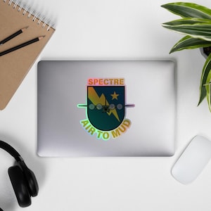 Op de afbeelding: Een holografische sticker met een groen, geel en zwart ontwerp. De sticker toont een vliegtuig met de tekst "SPECTRE AIR TO MUD" eromheen.