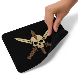 May include: Black rectangular mousepad featuring a skull with a feather and crossed swords and knives in tan and brown. The mousepad is being held up by a hand, revealing a black, textured underside.