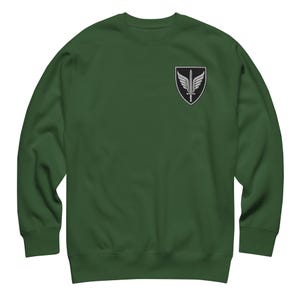 May include: A forest green crewneck sweatshirt featuring a black shield-shaped patch on the upper left chest. The patch displays a white graphic of a sword pointing downward, flanked by two symmetrical wings, all outlined in a thin white border.