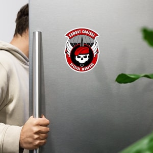 以下が含まれることがあります： 赤いベレー帽をかぶった頭蓋骨、翼、パラシュートのデザインが施された丸いマグネット。マグネットには「COMBAT CONTROL」と「SPECIAL WARFARE」の文字が赤と白で書かれています。