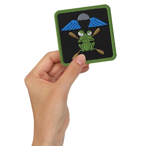 Peut inclure: Une grenouille verte portant un parachute avec des ailes bleues, tenant deux pagaies croisées. La grenouille est sur un fond noir avec une bordure verte.