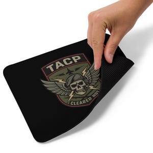 Pode incluir: Tapete de rato retangular preto com um gráfico de tema militar. O design inclui uma caveira usando boina e fone de ouvido, asas, raios e o texto "TACP CLEARED HOT". O tapete de rato é segurado por uma mão.