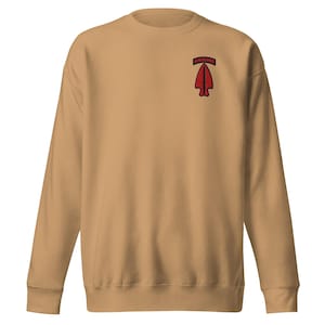 Pode incluir: Um moletom bege com gola redonda. Possui um patch vermelho em forma de ponta de flecha com a palavra "AIRBORNE" acima, localizado na parte superior esquerda do peito. O moletom é feito de um material macio. O design é simples e casual.