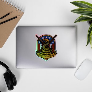May include: A sticker featuring a coiled yellow snake with a red tongue, set against a shield with crossed rifles and stars. The text "DON'T TREAD ON ME" is displayed below the snake in a banner. The sticker has a rainbow holographic effect.