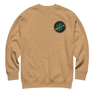 May include: A tan crewneck sweatshirt featuring a circular patch on the upper left chest. The patch displays two green footprints, a vertical silver dagger, and a horizontal yellow lightning bolt set against a black background with a gray border.