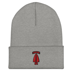 May include: A grey beanie hat with a turned-up brim. It has a red arrowhead patch with the word "AIRBORNE" embroidered above it. The hat is made of a soft, knitted material.