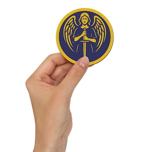 May include: A circular coaster with a detailed angel illustration holding a sword. The design is in yellow and gold on a navy blue background, with a yellow border. The coaster is held in a hand against a white backdrop.
