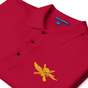 May include: A red polo shirt with a three-button placket and a collar. The shirt features a gold embroidered emblem of a skull with wings, a parachute, and a knife. The tag reads "PORT AUTHORITY".