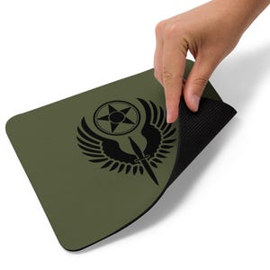 May include: Olive green rectangular mousepad featuring a black emblem with a star, wings, and a dagger. The mousepad has a black, textured underside. The image shows a hand lifting the edge of the mousepad.