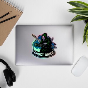 Puede incluir: Una pegatina holográfica con un personaje de la muerte con una capa con capucha negra, sosteniendo un arma. La pegatina tiene las palabras "GHOST RIDER" en una fuente estilizada, con un fondo azul y verde.