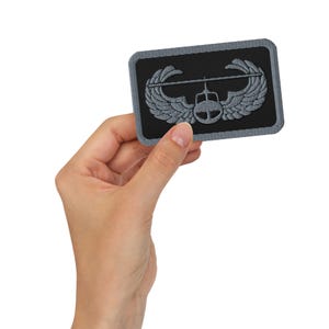 May include: A rectangular embroidered patch featuring a black background with a gray border. The design depicts a gray helicopter icon centered between two stylized wings, creating a symmetrical emblem with detailed stitching throughout.