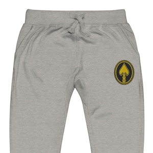 Pode incluir: Calças de moletom cinza claro com cintura com cordão e bolsos. Um emblema dourado e preto com um design de espadas está na perna esquerda. As calças de moletom são feitas de um material macio e confortável.