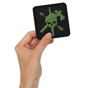 Peut inclure: Un écusson brodé noir avec un motif de crâne et d'os croisés verts. Le crâne porte un béret vert et est entouré d'armes croisées, dont une lance, une hache et une flèche.