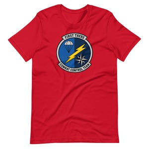 May include: A red short-sleeve t-shirt featuring a circular emblem on the chest. The emblem displays a parachute, a yellow lightning bolt, and a compass star, with the text FIRST THERE and COMBAT CONTROL TEAM printed along the border.