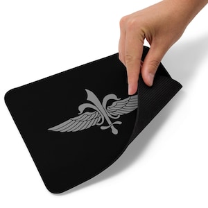 May include: Black rectangular mousepad featuring a silver emblem with wings and a fleur-de-lis design. The mousepad has a textured underside and is being held up by a hand.