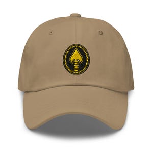 May include: A tan baseball cap with a curved brim. The cap features a circular embroidered patch with a gold spade design on a black background, surrounded by a gold rope-like border. The cap has a structured fit and a top button.