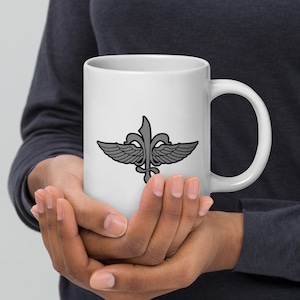 May include: White ceramic mug featuring a gray emblem with wings and a fleur-de-lis design. The mug is held by a person. The design is centered on the mug, offering a clean and modern aesthetic. The mug has a curved handle.