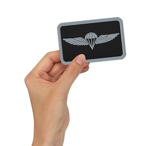 May include: A gray and black embroidered patch with a parachute and wings. The patch is surrounded by a gray border.