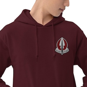 Pode incluir: Um moletom vinho com capuz e cordão. O patch no peito apresenta uma seta branca, asas e as palavras "VOLARE OPTIMOS". O moletom é feito de um material macio.