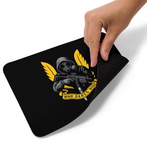 Op de afbeelding: Zwart rechthoekig muismat met een afbeelding van een figuur met een gasmasker die een geweer vasthoudt, met gele vleugels en de tekst "WHO DARES WINS" op een banner. Een hand tilt de muismat op.