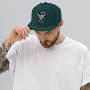 May include: A dark teal snapback cap featuring a centered embroidered patch. The patch is a black shield shape with a white border, containing a white graphic of a vertical sword flanked by two symmetrical wings, all rendered in clean white thread.