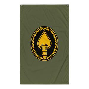 Pode incluir: Bandeira verde-oliva com um emblema de espadas dourado dentro de um oval preto, emoldurado por uma borda semelhante a uma corda. A espada tem um elemento em forma de escada na base. A bandeira é retangular e pendurada verticalmente.
