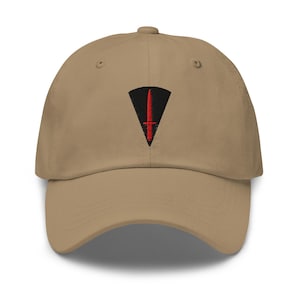 May include: A tan baseball cap featuring an embroidered patch on the front. The patch consists of a black triangular shape with a red dagger symbol centred within it. The cap has a curved brim and a structured crown with matching tan stitching.