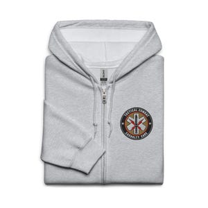 Könnte beinhalten: Ein hellgrauer Kapuzenpullover mit Reißverschluss und einem kleinen gestickten Aufnäher auf der Brust. Der Aufnäher zeigt ein Star-of-Life-Symbol mit den Worten "TACTICAL COMBAT CASUALTY CARE" in einem kreisförmigen Design.