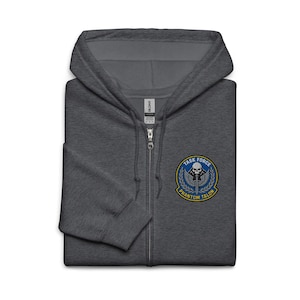 Könnte beinhalten: Ein dunkelgrauer Kapuzenpullover mit Reißverschluss, Kapuze und Frontreißverschluss. Der Hoodie hat einen runden Aufnäher auf der Brust mit den Worten "TASK FORCE PHANTOM TALON" um ein Totenkopf- und Schwertdesign. Der Hoodie ist aus weichem Material.