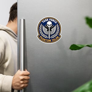 Puede incluir: Un parche circular con el texto "TASK FORCE" y "PHANTOM TALON" en letras doradas. El diseño presenta una calavera, una espada y alas, sobre un fondo azul con detalles dorados y blancos. El parche está adherido a una superficie metálica.