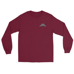 Peut inclure: Un t-shirt à manches longues bordeaux avec un petit motif gris détaillé sur le haut de la poitrine gauche. Le motif semble être un emblème stylisé. Le t-shirt a un col rond et des manches longues.