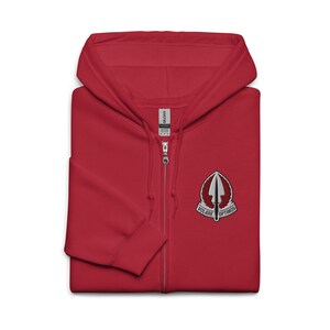 Pode incluir: Um moletom vermelho com capuz e zíper com um patch bordado em branco e preto no peito. O patch apresenta uma seta estilizada e as palavras "VOLARE OPTIMOS". O moletom tem capuz e zíper completo.