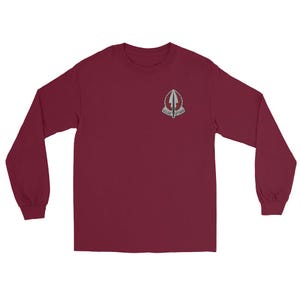 Pode incluir: Uma camisa de manga comprida bordô com um pequeno emblema na parte superior esquerda do peito. O emblema é um design prateado e branco com texto. A camisa tem gola redonda e mangas compridas.